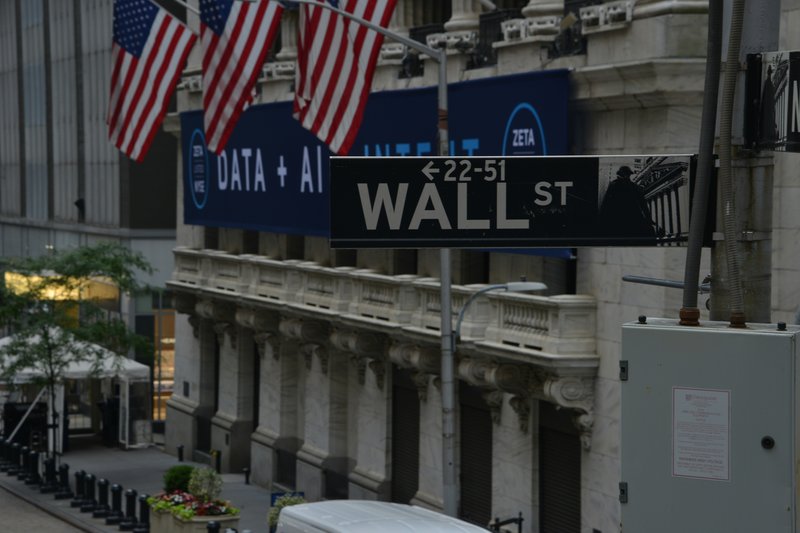 Fotografía de Wall Street, con letrero y banderas de EE.UU. en un entorno urbano financiero. Ilustra alternativas de inversión en EE.UU. con tasas del 10-20%. Mercurium Investech transforma vidas mediante literacy financiera, interés compuesto y manejo de portafolios innovador para libertad financiera.